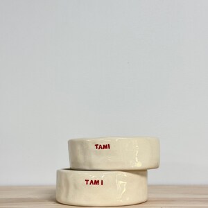 May include: Two stacked, off-white ceramic bowls with a slightly irregular, hand-formed texture. Each bowl features the name TAMI stamped in bold, red, uppercase letters on the front exterior surface, creating a simple and functional design.