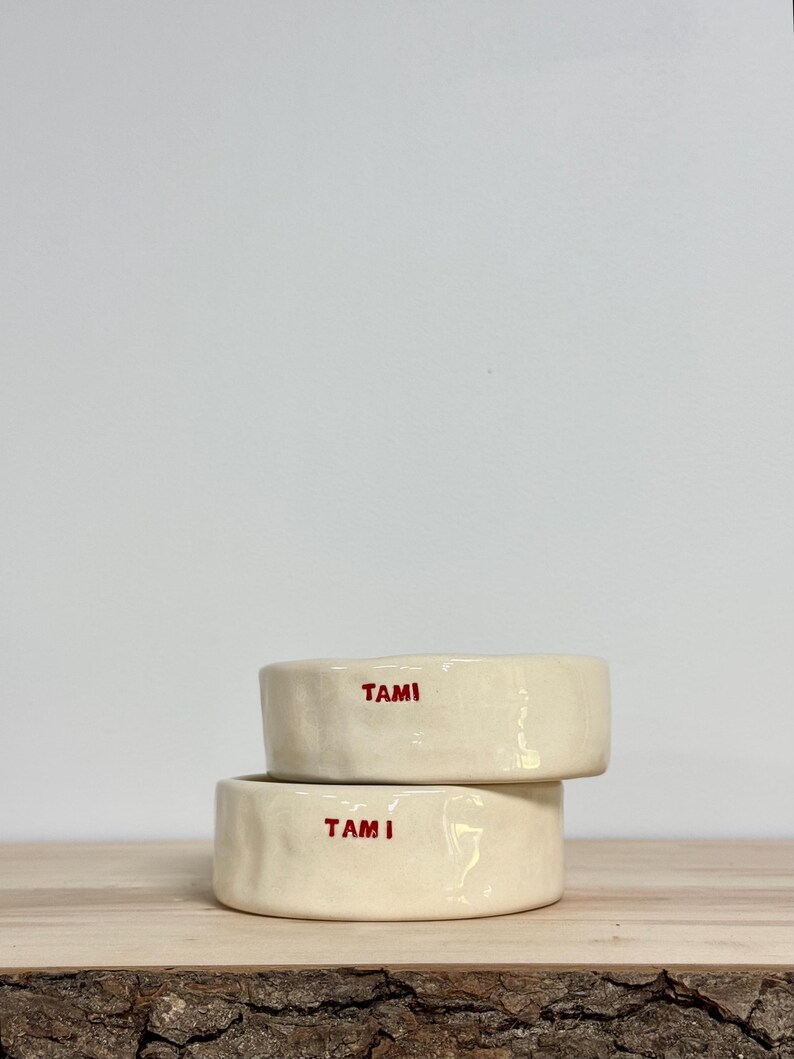 May include: Two stacked, off-white ceramic bowls with a slightly irregular, hand-formed texture. Each bowl features the name TAMI stamped in bold, red, uppercase letters on the front exterior surface, creating a simple and functional design.