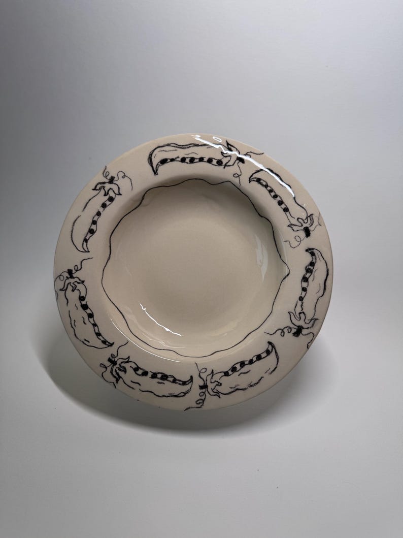 Puede incluir: Cuenco de cer&aacute;mica color crema con un dise&ntilde;o de vainas de guisantes negras alrededor del borde. El cuenco tiene un borde ligeramente ondulado y un interior poco profundo. El dise&ntilde;o de las vainas de guisantes est&aacute; dibujado a mano.