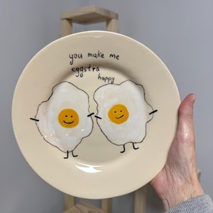 May include: A round, cream-coloured ceramic plate featuring a hand-painted illustration of two fried eggs with smiling faces, stick arms, and legs. The text "you make me eggstra happy" is written in black ink above the two egg characters.