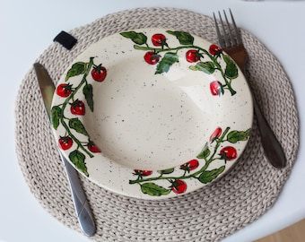 May include: A cream-colored ceramic plate with a hand-painted tomato vine design, set on a beige woven placemat. A stainless steel knife and a wooden fork are placed on either side of the plate. The plate is approximately 9 inches in diameter.