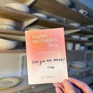 May include: A greeting card with a gradient pink and white background reads "HAPPY MOTHER'S Day". Handwritten text says "Love you soo much! Lisa" and "by V. Martin". The card is held in front of shelves with pottery.