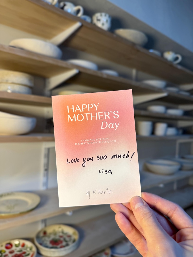 May include: A greeting card with a gradient pink and white background reads "HAPPY MOTHER'S Day". Handwritten text says "Love you soo much! Lisa" and "by V. Martin". The card is held in front of shelves with pottery.