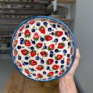 Pode incluir: Uma tigela de cerâmica redonda com interior na cor creme, apresentando um padrão repetitivo de morangos vermelhos com folhas verdes e pequenos mirtilos azul-escuros. A tigela tem uma borda espessa e salpicada em azul-escuro.