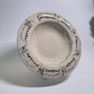 Puede incluir: Cuenco de cer&aacute;mica blanquecina con un dise&ntilde;o de vainas de guisantes negras dibujadas a mano alrededor del borde. El cuenco tiene una forma poco profunda y redondeada y un acabado brillante. El dise&ntilde;o de vainas de guisantes a&ntilde;ade un toque caprichoso.
