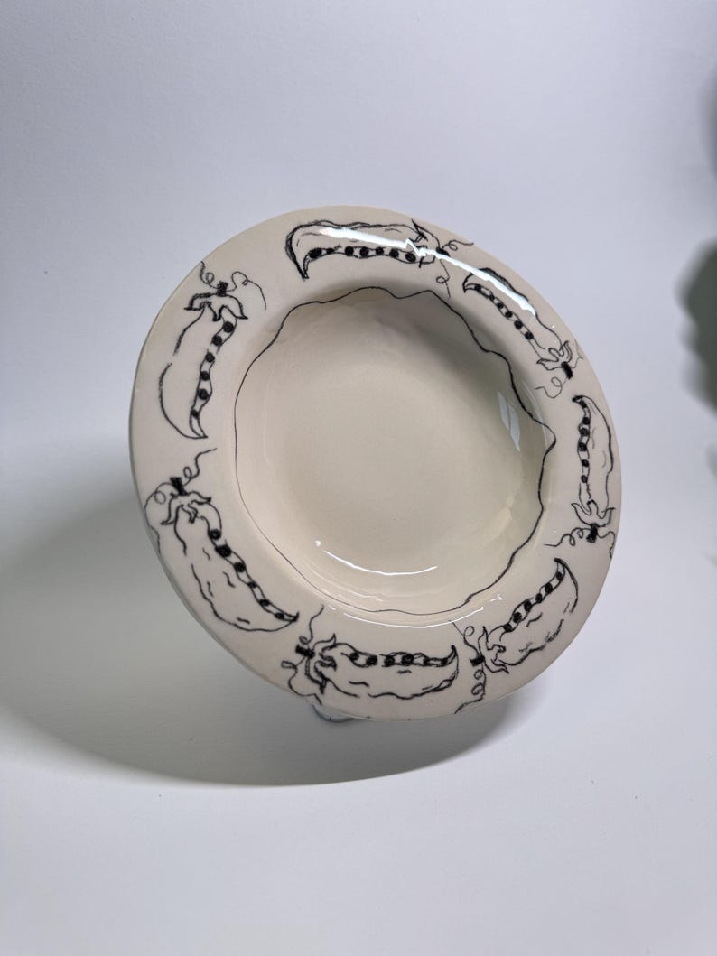 Puede incluir: Cuenco de cer&aacute;mica blanquecina con un dise&ntilde;o de vainas de guisantes negras dibujadas a mano alrededor del borde. El cuenco tiene una forma poco profunda y redondeada y un acabado brillante. El dise&ntilde;o de vainas de guisantes a&ntilde;ade un toque caprichoso.