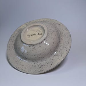 Puede incluir: Un cuenco de cer&aacute;mica con exterior gris moteado e interior color crema. La parte inferior del cuenco presenta el texto "by V. Martin" y un peque&ntilde;o logotipo. El cuenco est&aacute; ligeramente inclinado, mostrando su forma y dise&ntilde;o.