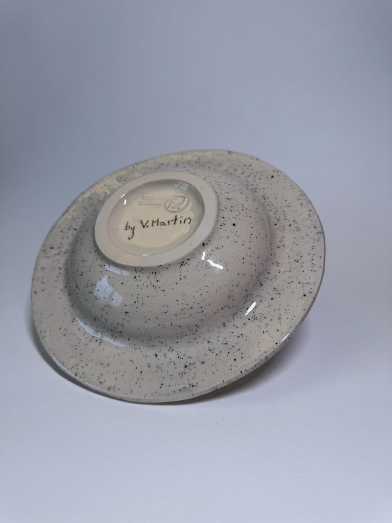 Puede incluir: Un cuenco de cer&aacute;mica con exterior gris moteado e interior color crema. La parte inferior del cuenco presenta el texto "by V. Martin" y un peque&ntilde;o logotipo. El cuenco est&aacute; ligeramente inclinado, mostrando su forma y dise&ntilde;o.