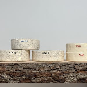 May include: Five ceramic bowls with a speckled off-white finish. Three bowls feature the black text COVIE, one bowl features the blue text DOMINO, and two bowls feature the red text TAMI. The bowls have a low, cylindrical shape with smooth edges.
