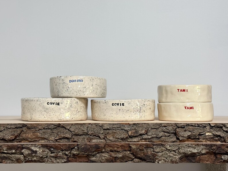May include: Five ceramic bowls with a speckled off-white finish. Three bowls feature the black text COVIE, one bowl features the blue text DOMINO, and two bowls feature the red text TAMI. The bowls have a low, cylindrical shape with smooth edges.