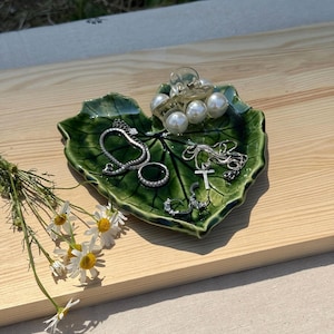 Può includere: Un piattino in ceramica verde lucido a forma di foglia, contenente un fermaglio per capelli trasparente con perle bianche, una collana a catena argentata con ciondolo a croce, un anello argentato e piccoli orecchini a cerchio, con margherite.