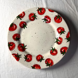 May include: A white ceramic plate decorated with hand-painted red tomatoes and green stems. The tomatoes are evenly spaced around the rim of the plate. The plate has a speckled background.