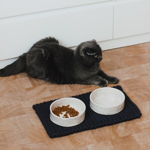 May include: Two round, white ceramic bowls with a speckled pattern placed on a rectangular, dark navy blue chunky knit mat. One bowl contains brown dry pet food kibble, while the other bowl is empty. The bowls have a smooth, cylindrical shape.