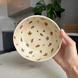 Puede incluir: Un cuenco redondo de cerámica con una base de color crema que presenta una textura moteada y un patrón de pequeñas hojas de otoño pintadas a mano en tonos naranja, amarillo y marrón, dispersas por la superficie interior y el borde.