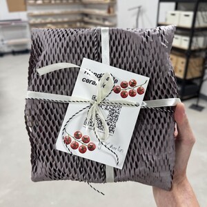 Puede incluir: Un paquete envuelto en papel de nido de abeja gris, atado con cinta blanca y cordel blanco y negro. Se adjunta una etiqueta blanca con ilustraciones de cerezas rojas y texto. El paquete lo sostiene una persona.