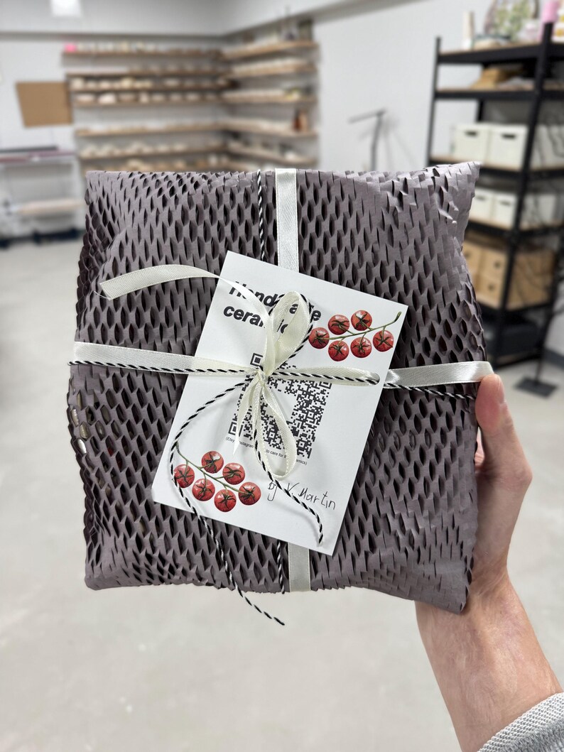 Puede incluir: Un paquete envuelto en papel de nido de abeja gris, atado con cinta blanca y cordel blanco y negro. Se adjunta una etiqueta blanca con ilustraciones de cerezas rojas y texto. El paquete lo sostiene una persona.