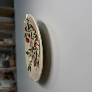 K&ouml;nnte beinhalten: Ein Keramikteller mit handgemaltem Design aus roten Tomaten und gr&uuml;nen Bl&auml;ttern auf wei&szlig;em Grund. Der Teller ist an einer hellgrauen Wand befestigt und unterstreicht seinen dekorativen Charakter. Der Teller hat einen einzigartigen, unregelm&auml;&szlig;igen Rand.