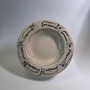 Puede incluir: Un cuenco de cer&aacute;mica color crema con borde festoneado. El borde est&aacute; decorado con un patr&oacute;n repetido de dibujos lineales negros de vainas de guisantes. El cuenco tiene poca profundidad y un acabado liso y brillante.