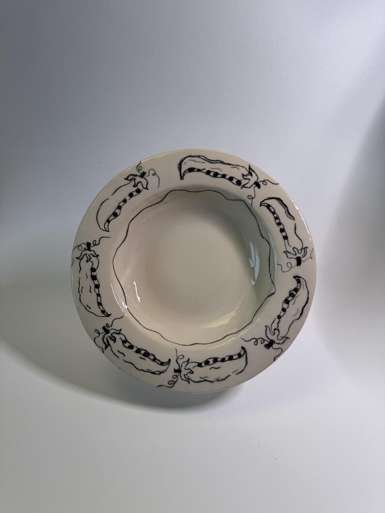 Puede incluir: Un cuenco de cer&aacute;mica color crema con borde festoneado. El borde est&aacute; decorado con un patr&oacute;n repetido de dibujos lineales negros de vainas de guisantes. El cuenco tiene poca profundidad y un acabado liso y brillante.