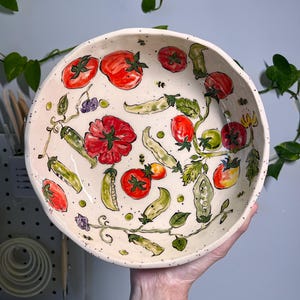 Puede incluir: Un cuenco de cerámica redondo y blanquecino con un diseño pintado a mano de tomates rojos, vainas de guisantes verdes y bayas moradas. El cuenco tiene una textura moteada y un borde ancho. La obra de arte es detallada y colorida.