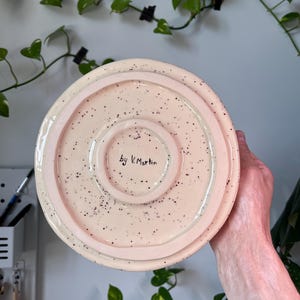 Op de afbeelding: Een keramische, gespikkelde, lichtroze plaat met een opstaande rand. De plaat heeft een glanzende afwerking en kleine zwarte spikkels. De tekst "by V. Martin" is met de hand geschreven op de bodem van de plaat. De plaat wordt door een persoon vastgehouden.
