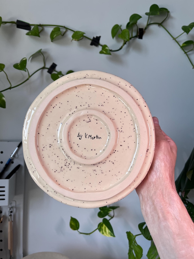 Op de afbeelding: Een keramische, gespikkelde, lichtroze plaat met een opstaande rand. De plaat heeft een glanzende afwerking en kleine zwarte spikkels. De tekst "by V. Martin" is met de hand geschreven op de bodem van de plaat. De plaat wordt door een persoon vastgehouden.
