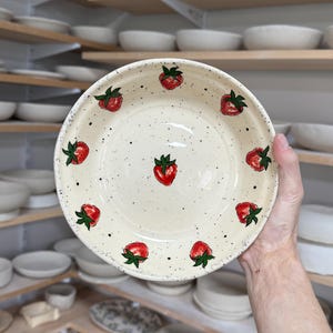 May include: A cream-coloured ceramic plate with a hand-painted strawberry design. The plate features red strawberries with green leaves and black speckles. The plate is held in front of a shelf of other ceramic items.