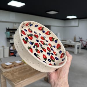 Op de afbeelding: Een keramische kom met een wit gespikkeld exterieur en een kleurrijk interieurontwerp. De binnenkant van de kom heeft een patroon van rode aardbeien en blauwe bosbessen. De kom is rond en heeft een licht verhoogde rand.