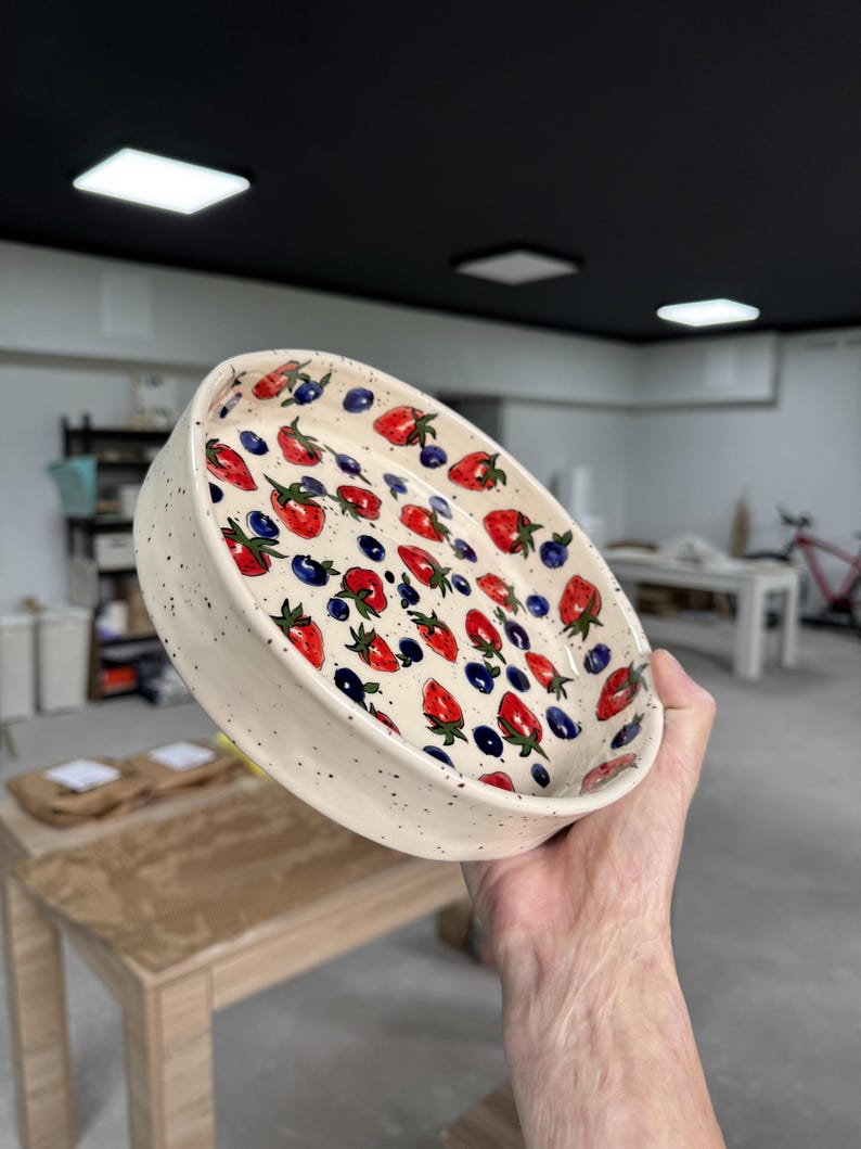 Op de afbeelding: Een keramische kom met een wit gespikkeld exterieur en een kleurrijk interieurontwerp. De binnenkant van de kom heeft een patroon van rode aardbeien en blauwe bosbessen. De kom is rond en heeft een licht verhoogde rand.