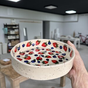 Op de afbeelding: Een ronde, cr&egrave;mekleurige keramische kom met een gespikkelde afwerking. De binnenkant is versierd met een patroon van rode aardbeien en blauwe bosbessen. De kom wordt in de hand van een persoon gehouden.