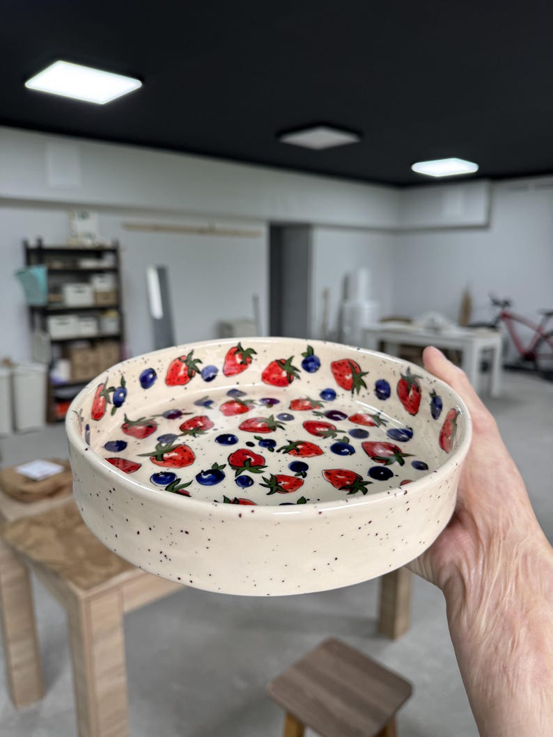 Op de afbeelding: Een ronde, cr&egrave;mekleurige keramische kom met een gespikkelde afwerking. De binnenkant is versierd met een patroon van rode aardbeien en blauwe bosbessen. De kom wordt in de hand van een persoon gehouden.