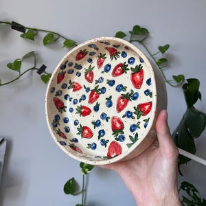 Op de afbeelding: Een ronde keramische kom met een cr&egrave;mekleurige, gespikkelde buitenkant. De binnenkant is versierd met een patroon van rode aardbeien, blauwe bosbessen en groene bladeren. De kom wordt door een hand omhoog gehouden.
