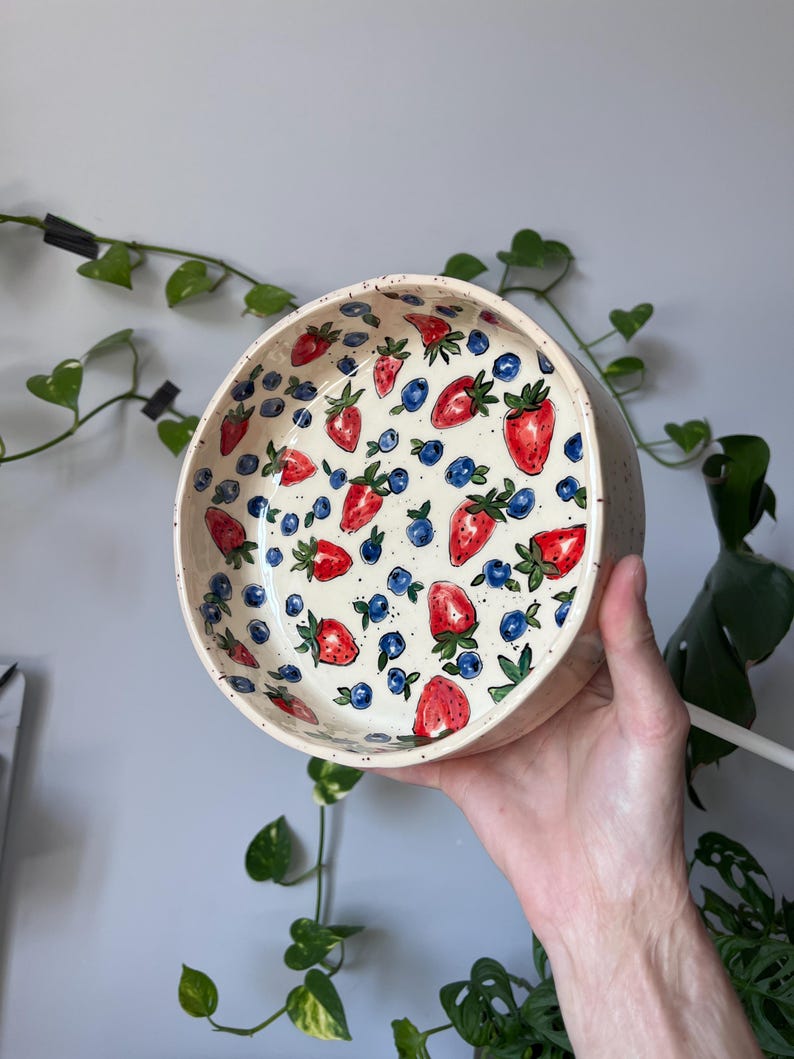 Op de afbeelding: Een ronde keramische kom met een cr&egrave;mekleurige, gespikkelde buitenkant. De binnenkant is versierd met een patroon van rode aardbeien, blauwe bosbessen en groene bladeren. De kom wordt door een hand omhoog gehouden.