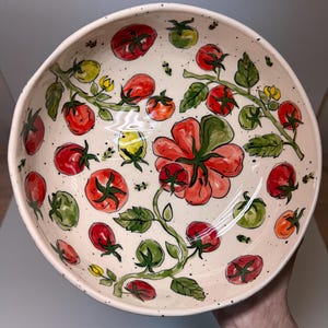 May include: A round ceramic bowl with a cream-colored base featuring a hand-painted pattern of red and green tomatoes on vines with small yellow flowers. The interior surface is speckled with black dots and shows a large central tomato blossom.