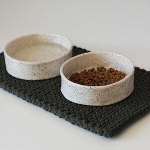 May include: Two round, speckled off-white ceramic bowls with low walls. One bowl is filled with water, and the other contains brown dry pet food. The bowls are placed on a dark green, rectangular, chunky knit fabric mat with a braided texture.