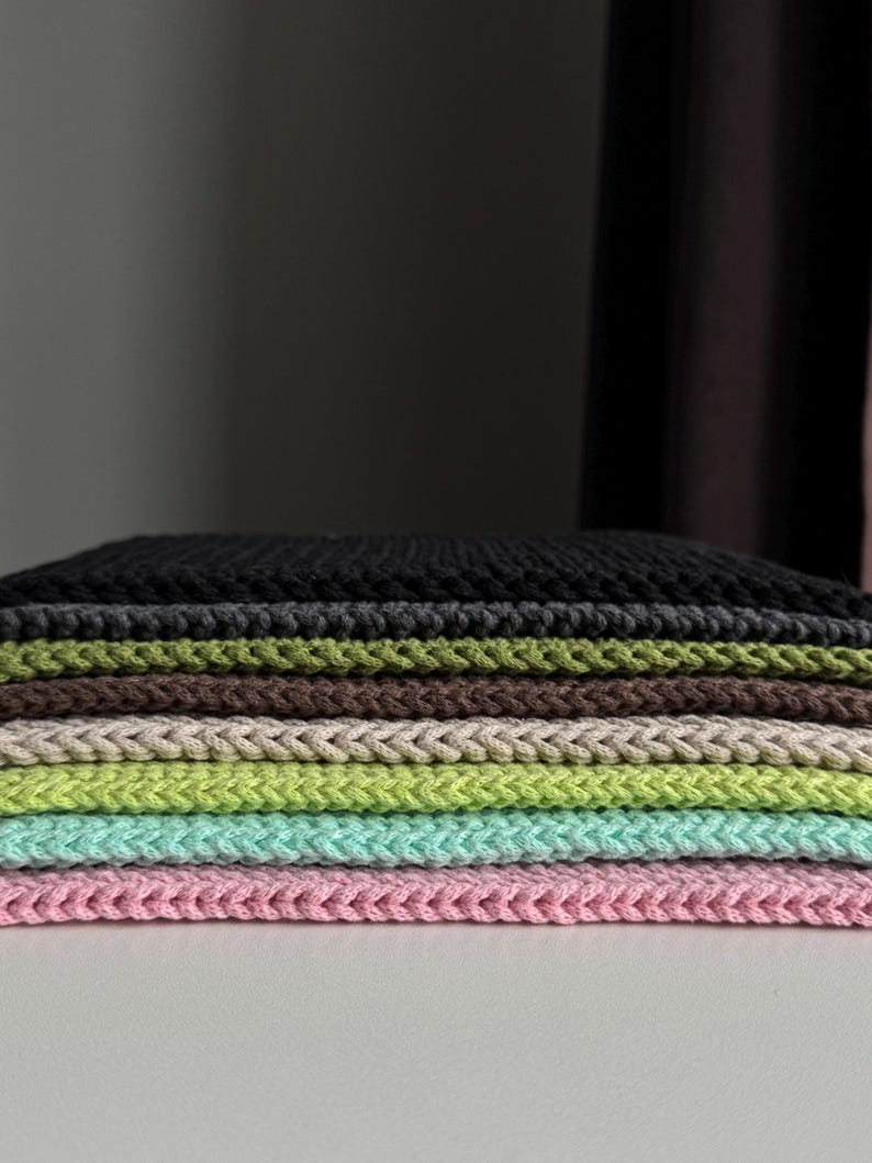 Puede incluir: Una pila de cuadrados de tela de punto coloridos. Los colores incluyen negro, verde oliva, marr&oacute;n, beige, verde claro, aguamarina y rosa. La tela tiene un aspecto texturizado y trenzado.