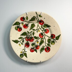 K&ouml;nnte beinhalten: Ein runder, cremefarbener Keramikteller mit einem handgemalten Tomatenranken-Design. Die Ranke hat rote und gr&uuml;ne Tomaten, gr&uuml;ne Bl&auml;tter und kleine gelbe Bl&uuml;ten. Der Teller hat eine gesprenkelte Textur.
