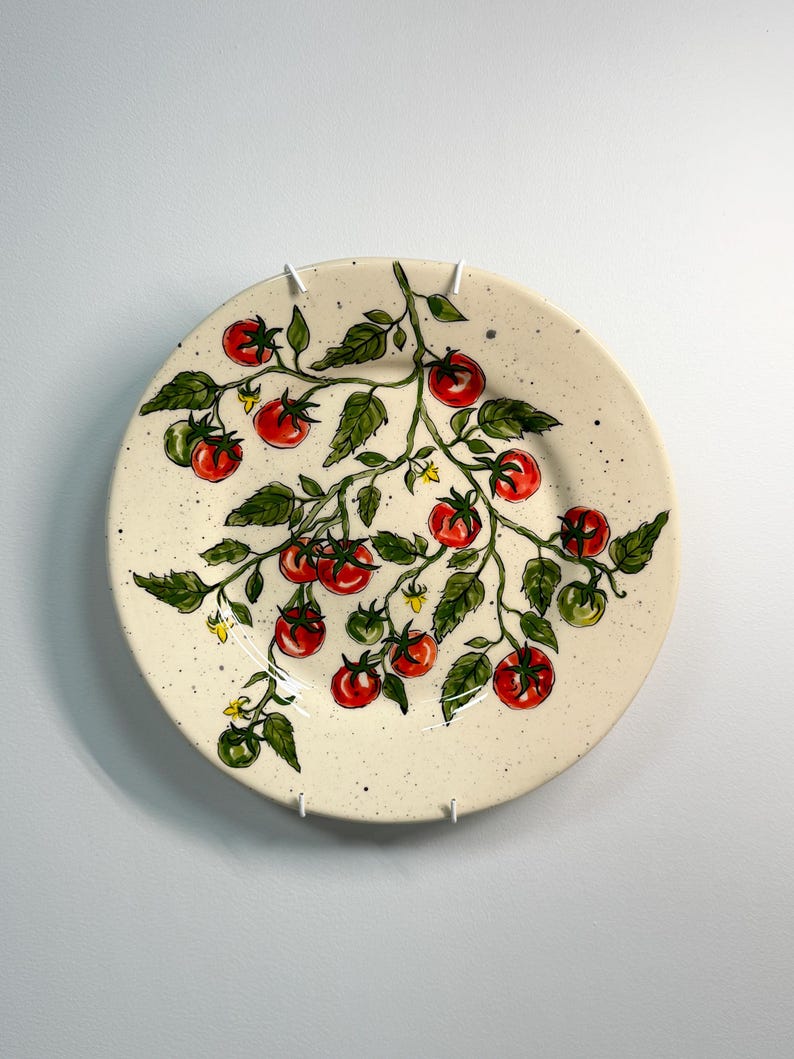 K&ouml;nnte beinhalten: Ein runder, cremefarbener Keramikteller mit einem handgemalten Tomatenranken-Design. Die Ranke hat rote und gr&uuml;ne Tomaten, gr&uuml;ne Bl&auml;tter und kleine gelbe Bl&uuml;ten. Der Teller hat eine gesprenkelte Textur.