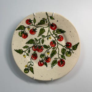 K&ouml;nnte beinhalten: Ein runder, cremefarbener Keramikteller mit handgemaltem Tomatenranken-Design. Die Ranke zeigt rote und gr&uuml;ne Tomaten, gr&uuml;ne Bl&auml;tter und kleine gelbe Bl&uuml;ten. Der Teller hat eine gesprenkelte Textur.