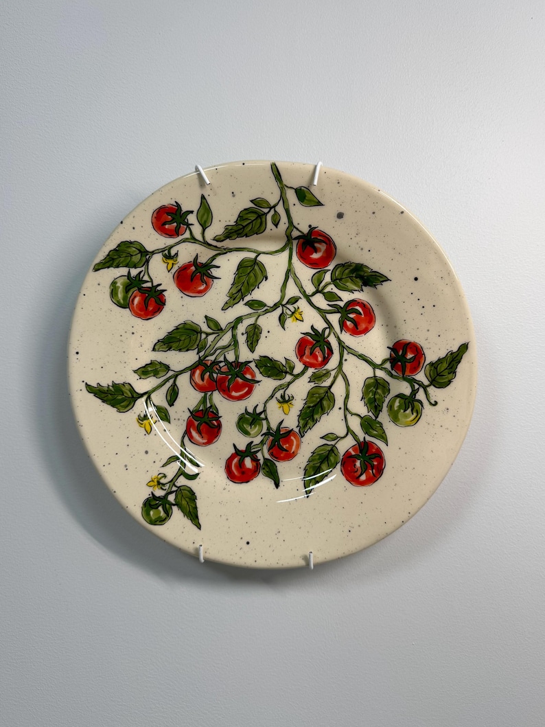 K&ouml;nnte beinhalten: Ein runder, cremefarbener Keramikteller mit handgemaltem Tomatenranken-Design. Die Ranke zeigt rote und gr&uuml;ne Tomaten, gr&uuml;ne Bl&auml;tter und kleine gelbe Bl&uuml;ten. Der Teller hat eine gesprenkelte Textur.