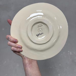 K&ouml;nnte beinhalten: Cremefarbene Keramikplatte mit erh&ouml;htem Rand, in der Hand gehalten. Die Unterseite zeigt einen zentralen, leicht erh&ouml;hten Kreis mit dem Text "MADE IN UKRAINE" und "by V. Martin". Die Platte hat eine gl&auml;nzende Oberfl&auml;che.