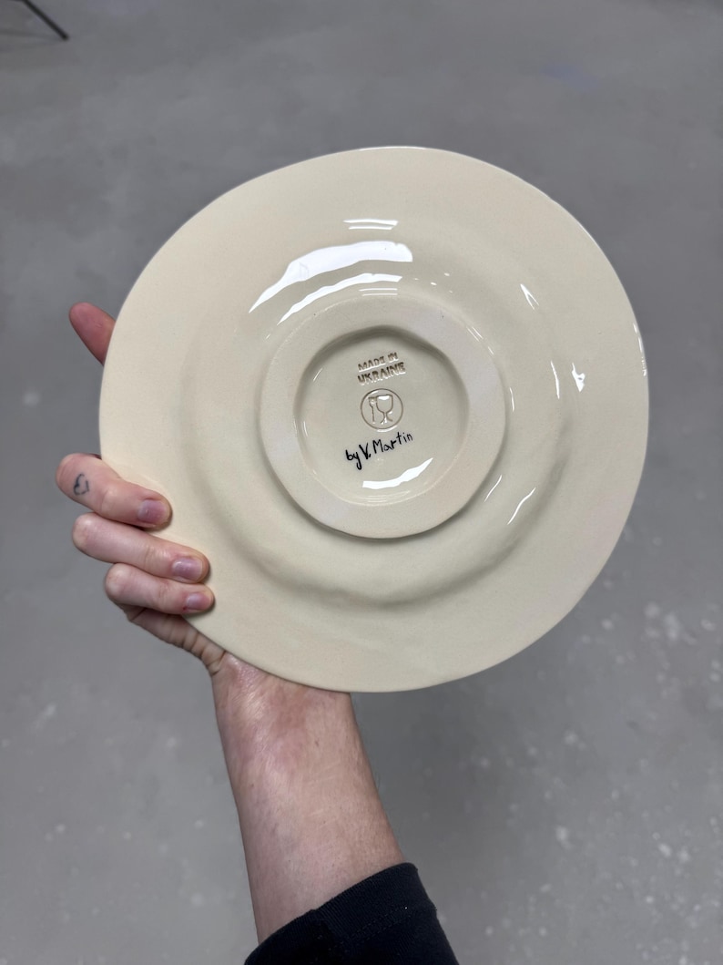 K&ouml;nnte beinhalten: Cremefarbene Keramikplatte mit erh&ouml;htem Rand, in der Hand gehalten. Die Unterseite zeigt einen zentralen, leicht erh&ouml;hten Kreis mit dem Text "MADE IN UKRAINE" und "by V. Martin". Die Platte hat eine gl&auml;nzende Oberfl&auml;che.