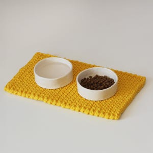 May include: A yellow crocheted pet food mat with two white ceramic bowls. One bowl contains water, and the other contains dry pet food. The mat is rectangular and has a textured surface, providing a comfortable and clean feeding area.