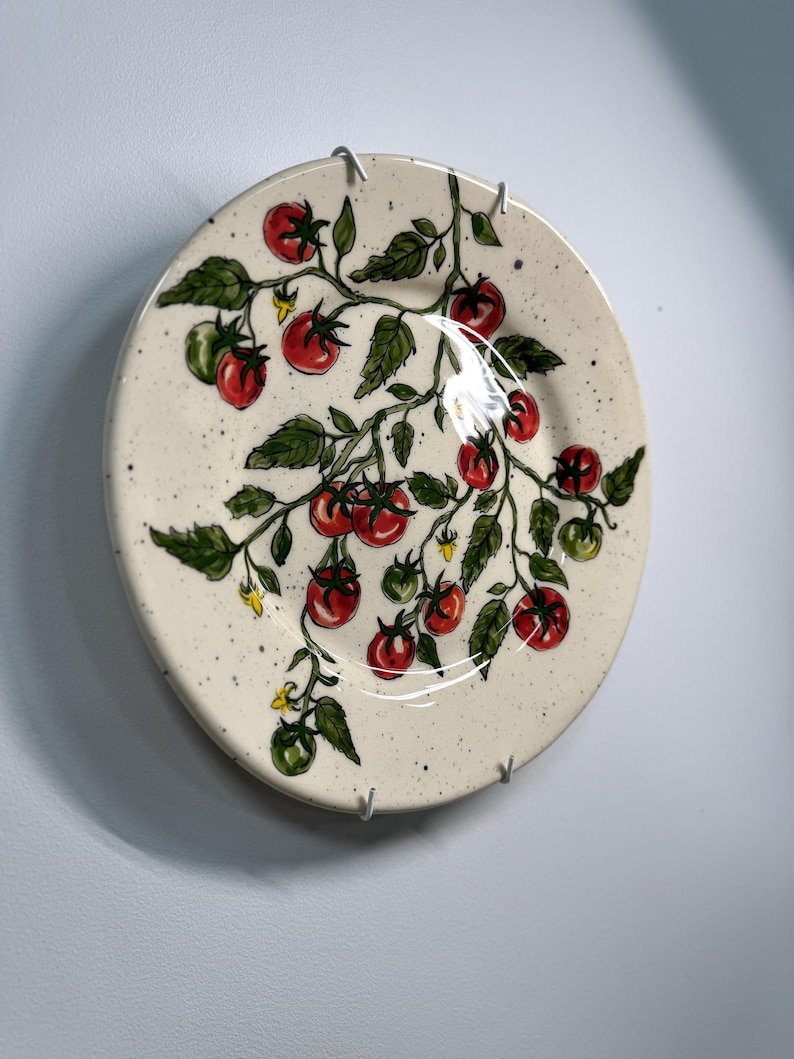 K&ouml;nnte beinhalten: Ein runder, cremefarbener Keramikteller mit einem handgemalten Tomatenranken-Design. Die Ranke zeigt rote und gr&uuml;ne Tomaten, gr&uuml;ne Bl&auml;tter und kleine gelbe Bl&uuml;ten. Der Teller ist gesprenkelt und mit Metallclips an der Wand befestigt.
