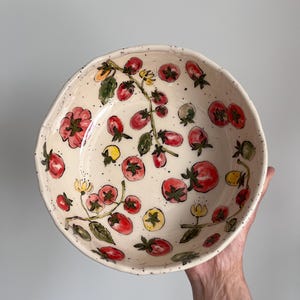 May include: A round ceramic bowl with an off-white base and a speckled finish. The interior features hand-painted illustrations of red and yellow tomatoes on green vines with small yellow flowers, distributed across the surface of the bowl.