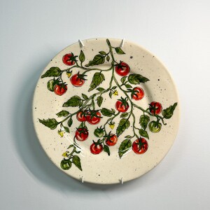 K&ouml;nnte beinhalten: Ein runder, cremefarbener Keramikteller mit einem handgemalten Design einer Tomatenranke. Die Ranke zeigt rote und gr&uuml;ne Tomaten, gr&uuml;ne Bl&auml;tter und kleine gelbe Bl&uuml;ten. Der Teller hat eine gesprenkelte Textur.