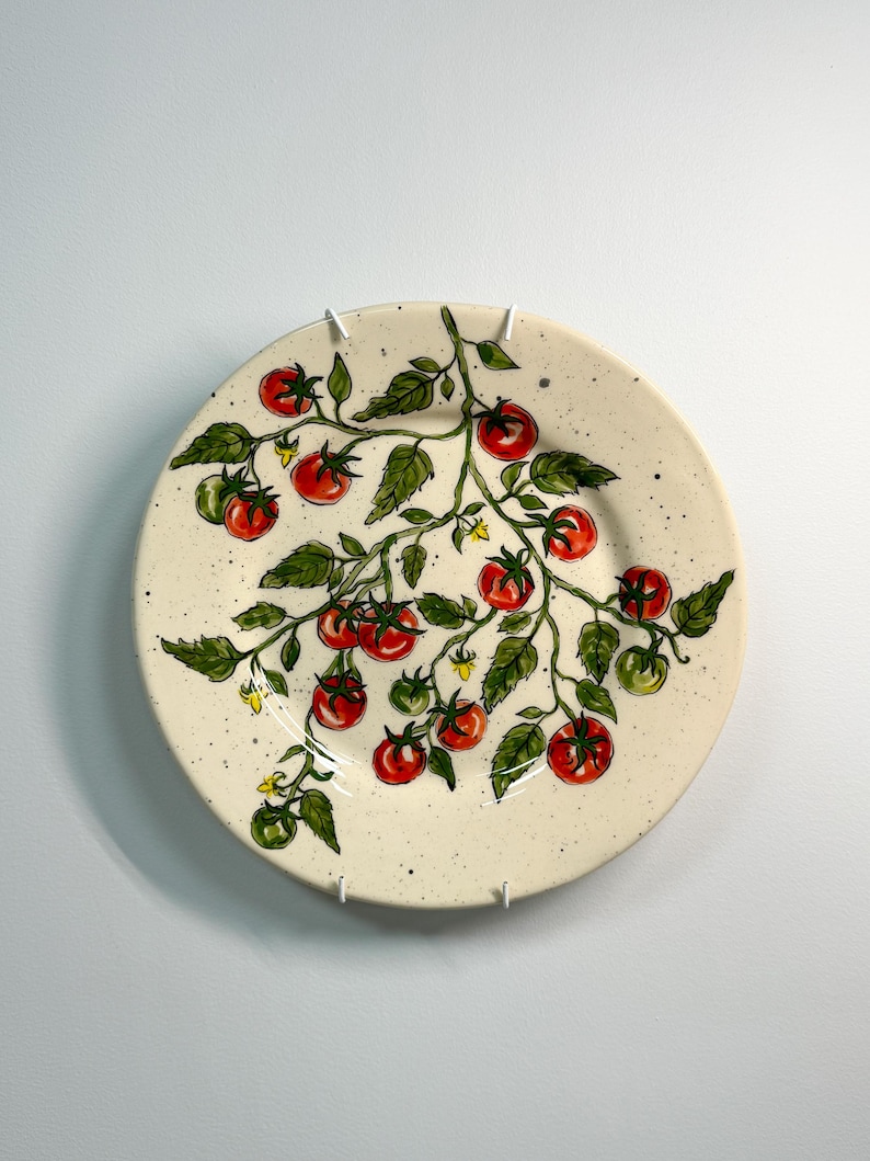 K&ouml;nnte beinhalten: Ein runder, cremefarbener Keramikteller mit einem handgemalten Design einer Tomatenranke. Die Ranke zeigt rote und gr&uuml;ne Tomaten, gr&uuml;ne Bl&auml;tter und kleine gelbe Bl&uuml;ten. Der Teller hat eine gesprenkelte Textur.
