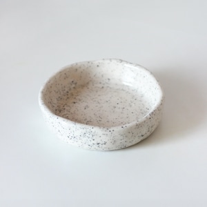 May include: A small, round ceramic dish with a slightly irregular, hand-formed rim. The surface is off-white with a speckled pattern of tiny black dots distributed across the interior and exterior, featuring a smooth, glossy finish throughout.