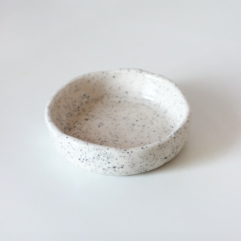May include: A small, round ceramic dish with a slightly irregular, hand-formed rim. The surface is off-white with a speckled pattern of tiny black dots distributed across the interior and exterior, featuring a smooth, glossy finish throughout.