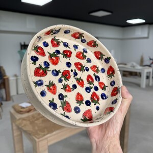 Op de afbeelding: Een ronde keramische kom met een cr&egrave;mekleurige, gespikkelde achtergrond. De kom is versierd met rode aardbeien en blauwe bosbessen. De aardbeien hebben groene steeltjes en bladeren. De kom wordt in de hand van een persoon gehouden.